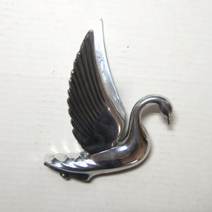 Può includere: Statuetta a forma di cigno color argento con ali spiegate. Il cigno ha un collo curvo e piume alari dettagliate. La statuetta è realizzata in metallo lucido e riflettente su sfondo bianco.