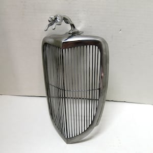 Könnte beinhalten: Ein verchromter Kühlergrill mit einem verchromten Hundefiguren-Kühlerfigur. Der Kühlergrill ist schildförmig und hat vertikale Streben. Der Hund springt mit offenem Maul nach vorne.