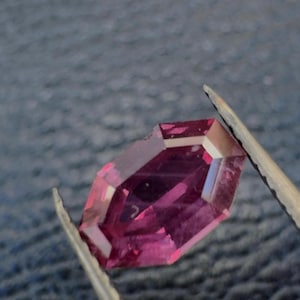 Peut inclure: Une pierre précieuse facettée et allongée, d'un magenta profond. La gemme est tenue par une pince, mettant en valeur sa taille géométrique et ses surfaces réfléchissantes. Les facettes captent la lumière.