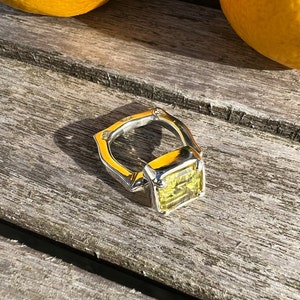 Peut inclure: Une bague en argent avec une grande pierre pr&eacute;cieuse carr&eacute;e jaune. La bague est pos&eacute;e sur une surface en bois.