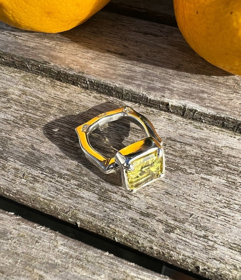 Peut inclure: Une bague en argent avec une grande pierre pr&eacute;cieuse carr&eacute;e jaune. La bague est pos&eacute;e sur une surface en bois.