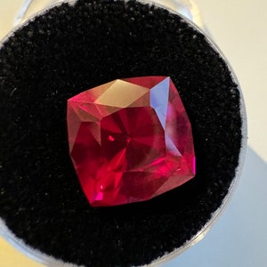 May include: A deep red gemstone with a square cut, set in a black velvet display case.