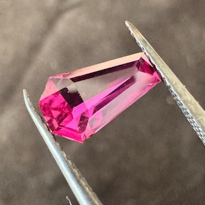 Peut inclure: Une pierre précieuse rose vif, taillée en rectangle, est maintenue par une pince argentée. La gemme transparente présente de multiples facettes qui reflètent la lumière. La pince est posée sur un fond gris neutre.