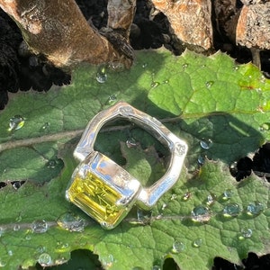 Peut inclure: Une bague en argent avec une grande pierre pr&eacute;cieuse jaune carr&eacute;e sertie au centre. La bague est pos&eacute;e sur une feuille verte avec des gouttelettes d'eau.