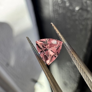 Könnte beinhalten: Ein facettierter, dreieckiger rosa Edelstein wird von einer Pinzette gehalten. Der Stein hat eine komplexe innere Struktur, die das Licht reflektiert. Die Pinzette ist silberfarben, der Hintergrund dunkel und unscharf.