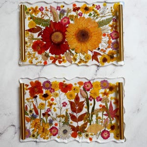 May include: Two rectangular clear resin trays with wavy edges and gold-toned metal handles. Each tray is embedded with a variety of pressed dried flowers and leaves in vibrant shades of red, orange, yellow, pink, and green, arranged in a dense floral pattern.