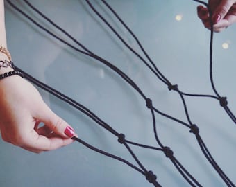 Peut inclure: Gros plan sur une main tenant un cordon de macramé noir, avec des nœuds formant un motif en forme de filet. La personne porte un bracelet doré, un ras-de-cou noir et un bracelet de perles noires. Fond bleu clair.