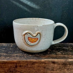 Könnte beinhalten: Eine gesprenkelte Keramiktasse mit einem Vogeldesign. Die Tasse ist cremefarben mit braunen Sprenkeln und einem hellblauen Vogel mit orangefarbenem Bauch. Die Tasse hat eine abgerundete Form und einen Henkel.