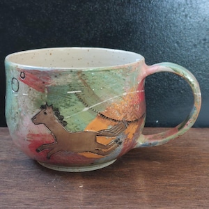 Könnte beinhalten: Ein Keramikbecher mit gesprenkeltem, cremeweißem Innenraum und einem Henkel. Die Außenseite zeigt ein handgemaltes abstraktes Design in Rosa-, Grün- und Orangetönen mit einer braunen Pferdeillustration und einer dünnen schwarzen Linie.