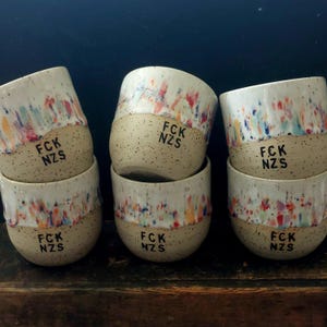 Könnte beinhalten: Ein Set aus fünf Keramikbechern mit gesprenkelter beiger Basis und einem farbenfrohen Tropfglasurmuster in Blau, Rot und Orange. Jeder Becher trägt den schwarzen Schriftzug FCK NZS im unteren Bereich. Die Becher sind in einer Gruppe von drei unten und zwei oben angeordnet.