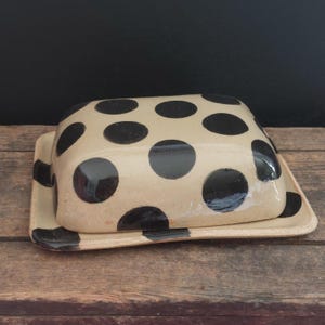 Könnte beinhalten: Eine Butterschale aus Keramik mit Deckel. Die Schale ist beige mit schwarzen Punkten. Der Deckel hat ein passendes Design.