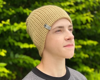 Puede incluir: Un gorro de punto beige con una pequeña etiqueta plateada que dice "#SBB".