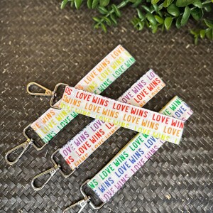 Op de afbeelding: Meerdere witte polsbandjes met de tekst "LOVE WINS" in regenboogkleuren. Elk sleutelhanger heeft een zilverkleurige sluiting. De sleutelhangers liggen op een donkerbruine, geweven ondergrond met groene vegetatie.