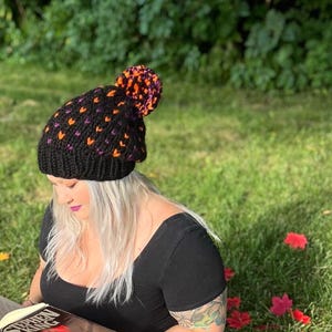 May include: A black knit beanie with a pom pom on top. The beanie has a pattern of orange and purple hearts. The beanie is worn by a person with blonde hair, who is reading a book.