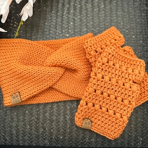 May include: A set of crocheted accessories in a warm orange color. The set includes a headband with a twisted design and a pair of fingerless gloves. Both pieces have a small brown leather tag with the text "SBB" on them.
