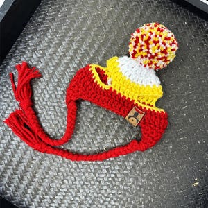K&ouml;nnte beinhalten: Rote, gelbe und wei&szlig;e H&auml;kelm&uuml;tze mit einem Bommel oben und einem langen roten Quast. Die M&uuml;tze ist aus dickem Garn gefertigt und hat ein schlichtes Design.
