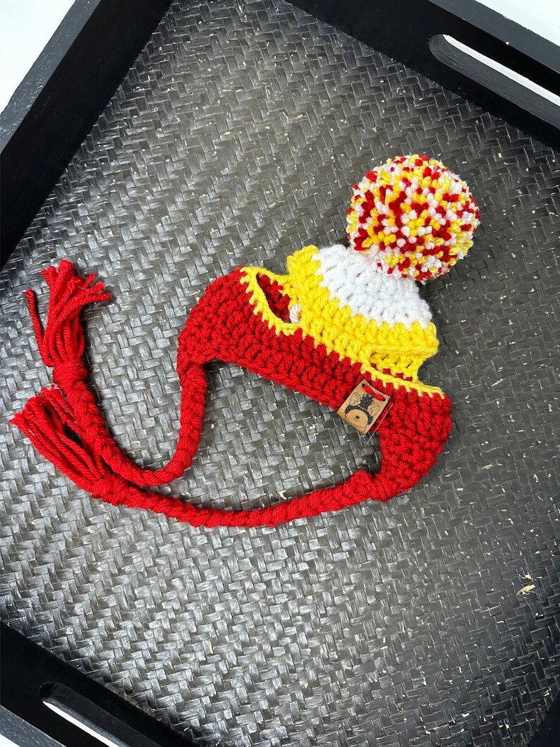 K&ouml;nnte beinhalten: Rote, gelbe und wei&szlig;e H&auml;kelm&uuml;tze mit einem Bommel oben und einem langen roten Quast. Die M&uuml;tze ist aus dickem Garn gefertigt und hat ein schlichtes Design.