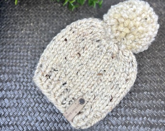 Może przedstawiać: Wełniany beret w kolorze kremowym z puszystym pomponem na górze. Beret ma brązową skórzaną metkę z napisem "Cozy Bunny Beanie".