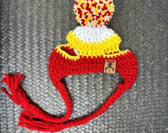 Puede incluir: Sombrero de perro de crochet rojo, blanco y amarillo con un pompón en la parte superior y una larga borla roja. El sombrero tiene una pequeña etiqueta marrón con una huella de pata blanca.