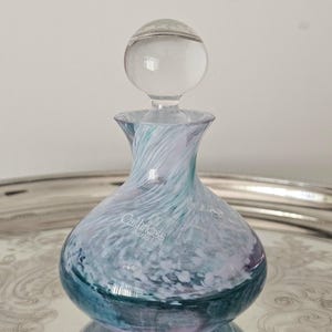 May include: A Caithness Scotland glass perfume bottle with a spherical clear glass stopper. The body features a swirling pattern of teal, white, and lavender colours. The brand name Caithness Scotland is printed in white on the front of the glass.