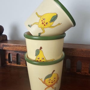 May include: Three stacked cream-coloured ceramic cups with dark green rims. Each cup features a different cartoon fruit character with a face, arms, and legs: a yellow lemon, a yellow pear, and an orange citrus fruit, all depicted in a playful pose.