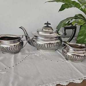 May include: A silver-plated tea set comprising a teapot, a sugar bowl and a creamer. Each piece features a rounded body with vertical fluted detailing along the lower half, polished metallic surfaces, and dark-coloured handles and finial.
