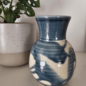 May include: A ceramic vase with a rounded body and a flared neck, featuring a glossy finish with a pattern of deep blue abstract shapes over a cream-coloured base. The surface shows subtle horizontal ridges from the pottery wheel process.