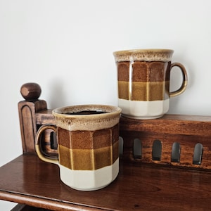 May include: Two ceramic mugs featuring a faceted, multi-sided design with horizontal bands of colour. The mugs display a gradient pattern transitioning from a dark brown top rim to a medium brown middle section and a cream-coloured base.