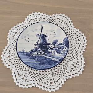May include: A round ceramic plate featuring a blue and white illustration of a traditional windmill and rural houses near a body of water, resting on a white, star-shaped, intricate crochet doily with a delicate, openwork lace pattern.