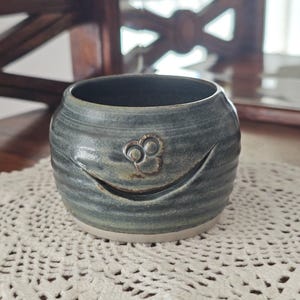 May include: A round, dark blue-grey ceramic yarn bowl with a smooth, glossy finish and horizontal ridges. The bowl features a curved cutout on the front side for yarn to pass through and a small, raised, circular decorative emblem above the opening.