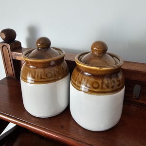 May include: Two ceramic storage jars with lids, featuring a two-tone design with a glossy brown upper section and a smooth white lower body. Each jar has the word NILONS embossed in the brown glaze on the neck, and a round knob on the lid.