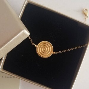May include: A gold chain bracelet with a circular pendant featuring a spiral design. The bracelet is displayed in a black box with a gold interior.