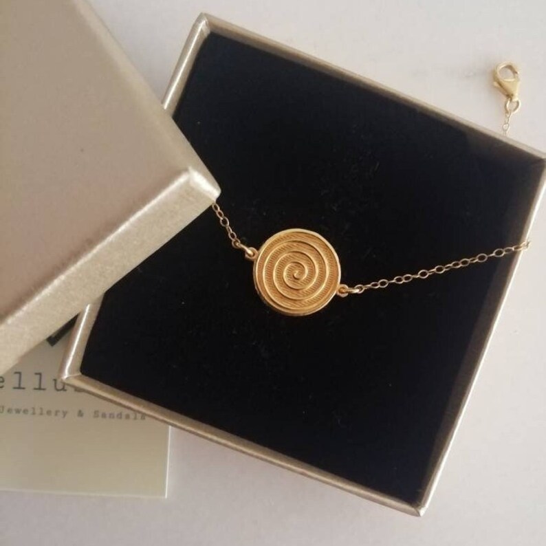 May include: A gold chain bracelet with a circular pendant featuring a spiral design. The bracelet is displayed in a black box with a gold interior.