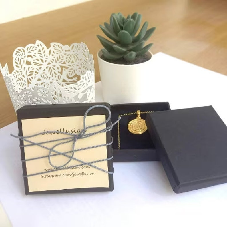 May include: A black gift box with a gold pendant necklace inside. The pendant is a circular design with a spiral pattern. The box is tied with a gray ribbon and has a small card with the text "Jewellusion" and website information.