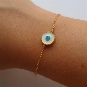 Puede incluir: Pulsera de cadena dorada con un dije de ojo turco blanco y azul. El dije tiene una base redonda de esmalte blanco con un ópalo azul en el centro.