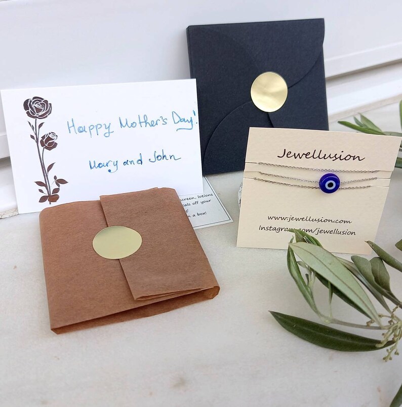 May include: A small gift box wrapped in brown paper with a gold sticker. A card with the text "Happy Mother's Day! Mary and John" is next to the box. A black envelope with a gold sticker is in the background. A small card with the text "Jewellusion" and a blue evil eye charm bracelet is in the foreground.