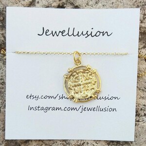 May include: A gold-colored pendant necklace featuring a circular coin-like charm with engraved text. The charm is suspended from a delicate chain. The background includes a white card with the text "Jewellusion".