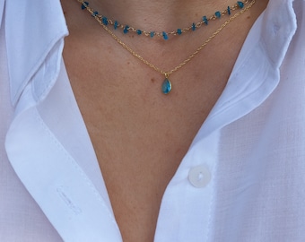 May include: A gold chain necklace with a teardrop-shaped blue gemstone pendant. The necklace is layered over a second gold chain necklace with small blue gemstone beads.