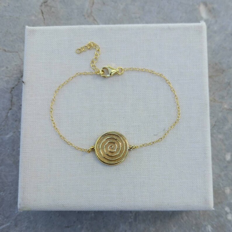 May include: A gold chain bracelet with a circular pendant featuring a spiral design.