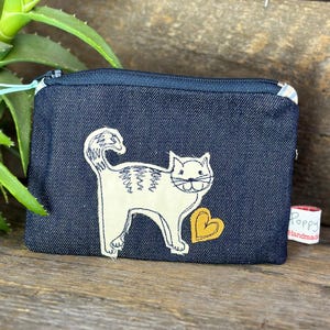 May include: A small denim zippered pouch with a white embroidered cat holding a yellow heart. The pouch has a tag that says "Poppy Handmade".