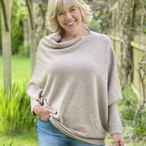 May include: A woman wearing a beige cowl neck sweater with long sleeves and blue jeans. She is standing in a garden setting.