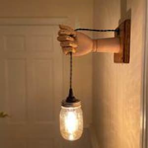 May include: A wall-mounted light fixture featuring a wooden hand holding a glass jar pendant light. The hand is painted a flesh tone and is attached to a wooden base. The jar has a textured surface and a metal lid, with a lit bulb inside.