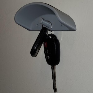 May include: A gray plastic key holder with a curved top and a hook for hanging keys. Two black car keys are hanging from the hook.