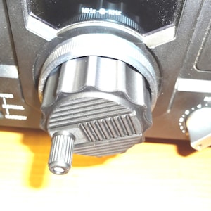 May include: A black electronic control knob with a textured, ridged surface and a smaller cylindrical adjustment dial attached to the front. The main dial features a grooved edge and is positioned on a black panel with the text MHz-kHz visible.