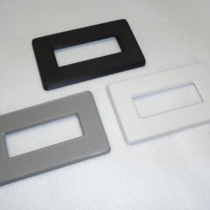 May include: Three wall plate covers in different colors: black, gray, and white. Each plate has a rectangular cutout in the center.
