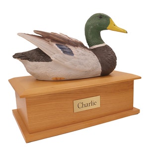 May include: A wooden pet urn with a realistic mallard duck figurine on top. The urn has a brass nameplate that reads "Charlie".