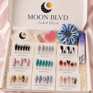 May include: A white gift box containing nine sets of press-on nails on branded cards, a light blue satin scrunchie, a small pink heart-shaped sponge, a nail file, and a purple tube of cuticle oil. The box lid features the Moon Blvd logo.