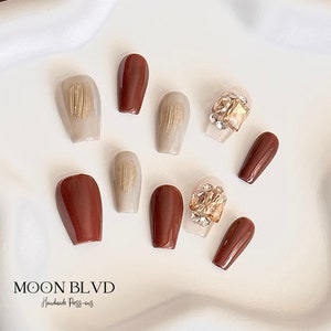 May include: Set of ten press-on nails with a brown and beige color scheme. Some nails feature a textured design, while others have gold and clear rhinestones.