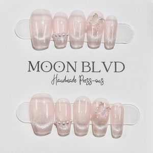 May include: Set of ten pink and white press-on nails with glitter and pearl accents. Some nails feature a butterfly charm. The nails are arranged on a white card with the text "MOON BLVD Handmade Press-ons".