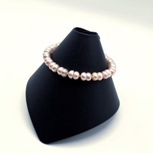 Puede incluir: Un brazalete de perlas rosa claro exhibido en un expositor de joyas negro. El brazalete está compuesto por numerosas perlas pequeñas y redondas ensartadas. Las perlas tienen un brillo suave y lustroso, sobre fondo blanco.